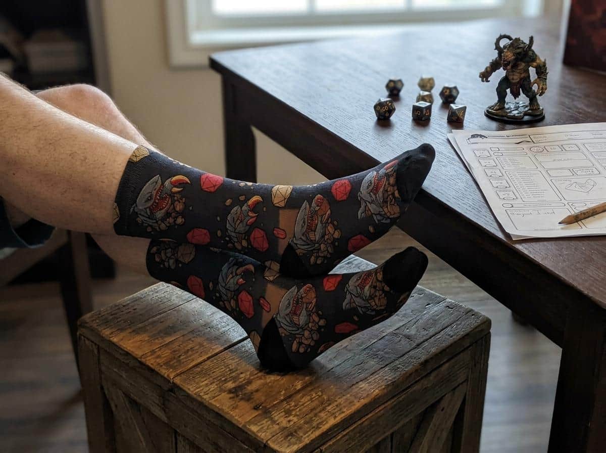 Photo 3 of 6: Humorous, combat-ready TTRPG crew socks featuring the notorious 'landshark' monster ambushing d20 dice in a seamless pattern, shown with feet up beside the game table